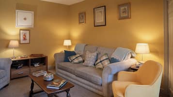 A living room with a sofa, armchairs, a coffee table, and lamps. There are framed pictures on the wall and a bookshelf with books.