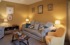 A living room with a sofa, armchairs, a coffee table, and lamps. There are framed pictures on the wall and a bookshelf with books. - Churchill Cottage (Bishop Auckland)