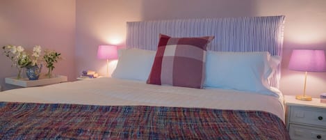 Una cama con colcha a cuadros, dos almohadas, una mesita de noche con una lámpara y un florero con flores.