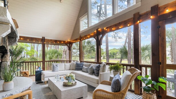 A covered outdoor seating area with a sofa, chairs, and tables, surrounded by large windows and wooden beams. - Charming cottage in popular neighborhood (Kiawah Island)