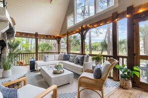 A covered outdoor seating area with a sofa, chairs, and tables, surrounded by large windows and wooden beams. - Charming cottage in popular neighborhood (Kiawah Island)