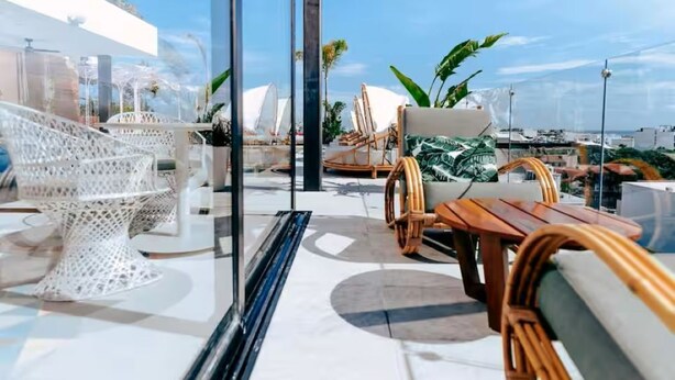 A rooftop terrace with wicker furniture, a glass balustrade, and a view of the cityscape. - Chic Queen Deluxe Suite in the Heart of PDC (Playa del Carmen)