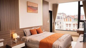 A modern hotel room with a bed, bedside table, lamp, and a view of buildings through a window. - Wake Up to Sunshine in Ninh Binh's Old Quarter - Cozy 2BR for Families! (Hoa Lư)