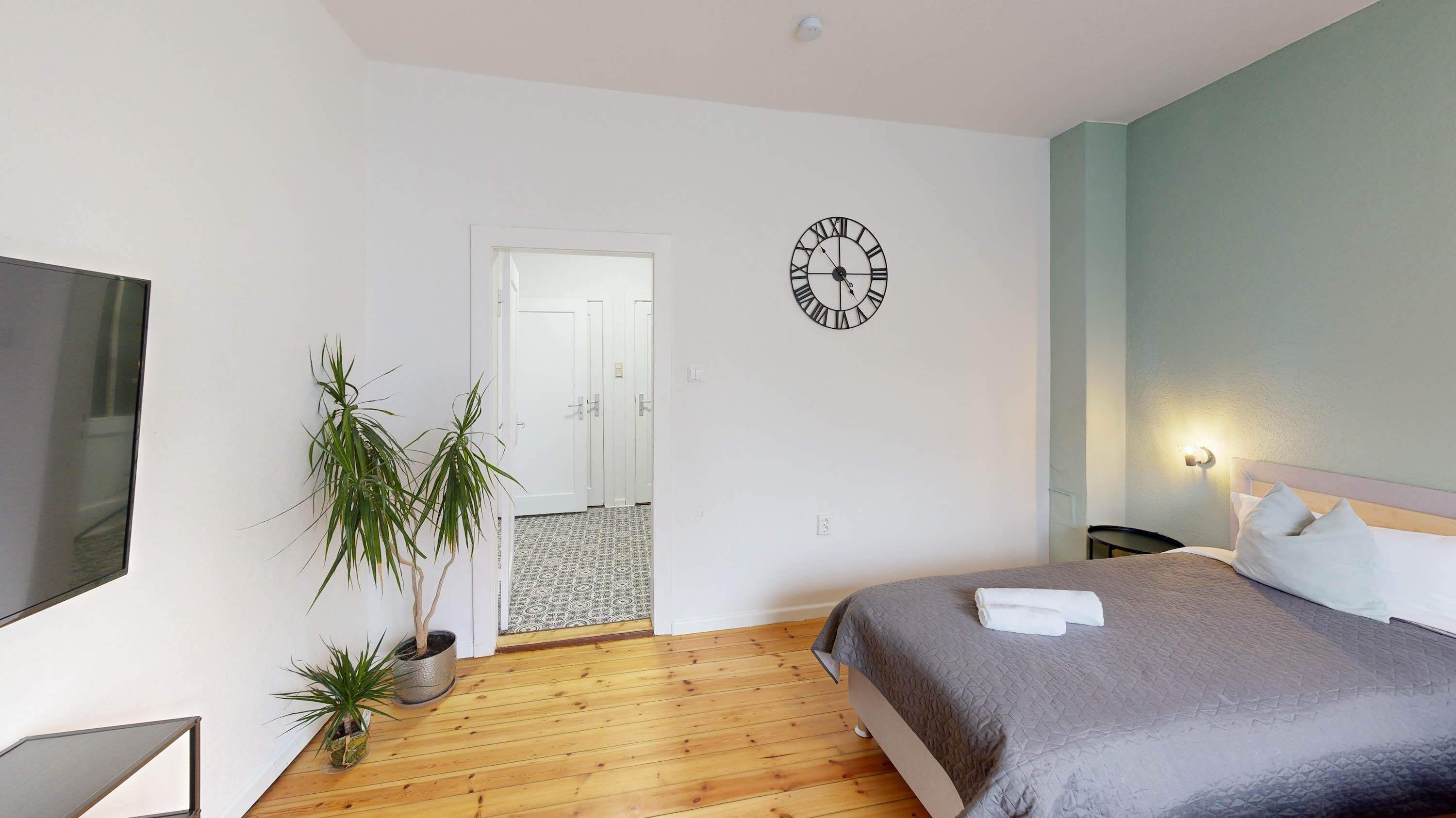 A bedroom with a bed, a wall clock, a TV, and a potted plant.