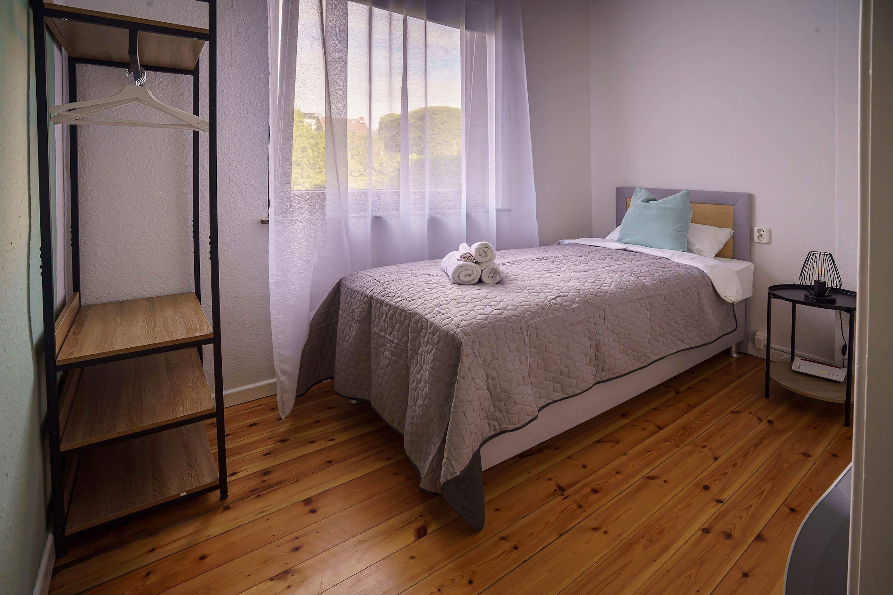 A bedroom with a bed, a bedside table, a hanging wardrobe, and a window with sheer curtains.
