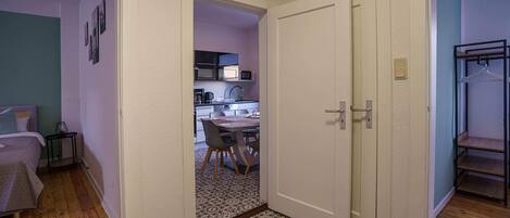 A hallway leading to a bedroom with a bed, a small table with a lamp, and a kitchenette area with a microwave and sink.
