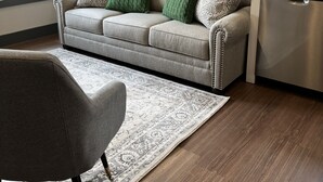 Ein modernes Wohnzimmer mit einem grauen Sofa, grünen Zierkissen, einem gemusterten Teppich, einem Essbereich mit einer Kaffeemaschine und einem Fenster mit Vorhängen.