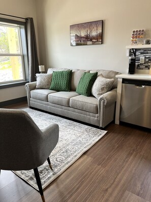 Un salon moderne avec un canapé gris, des coussins verts, un tapis à motifs, un coin repas avec une machine à café et une fenêtre avec des rideaux.