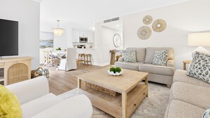Un salon moderne doté d’un téléviseur à écran plat, d’une table basse en bois, d’un canapé beige et d’un coin repas avec une cuisine en arrière-plan.