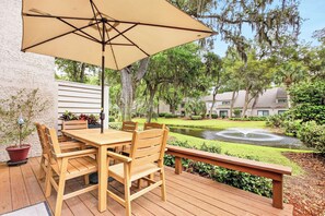 Una terraza de madera con una mesa y sillas, un gran paraguas y un estanque con una fuente.