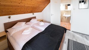 A bedroom with a wooden ceiling, a bed with white linens, a nightstand, a door leading to a bathroom with a sink. - VILLA LOTUS PANZIÓ (Zalakaros)