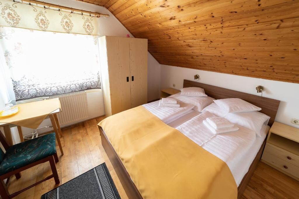 A bedroom with a wooden ceiling, a bed with white bedding, a wooden desk, and a chair.