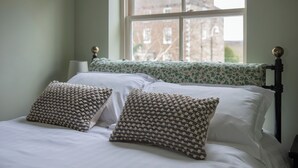 A bed with white linens and patterned pillows, a window with a view of buildings, and a nightstand with a lamp. - Sheriff's Tower Cottage - West (Bishop Auckland)