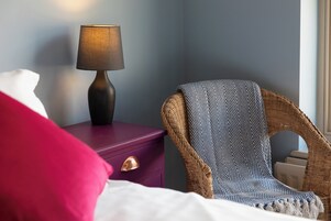 Una habitación con una cama, una mesita de noche con lámpara, una silla de mimbre y un plaid estampado.
