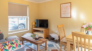 A living room with a TV, wooden furniture, a window with blinds, a framed picture on the wall, and a vase with flowers. - Sheriff's Tower Cottage - West (Bishop Auckland)
