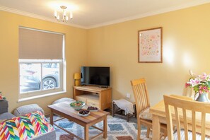A living room with a TV, wooden furniture, a window with blinds, a framed picture on the wall, and a vase with flowers. - Sheriff's Tower Cottage - West (Bishop Auckland)