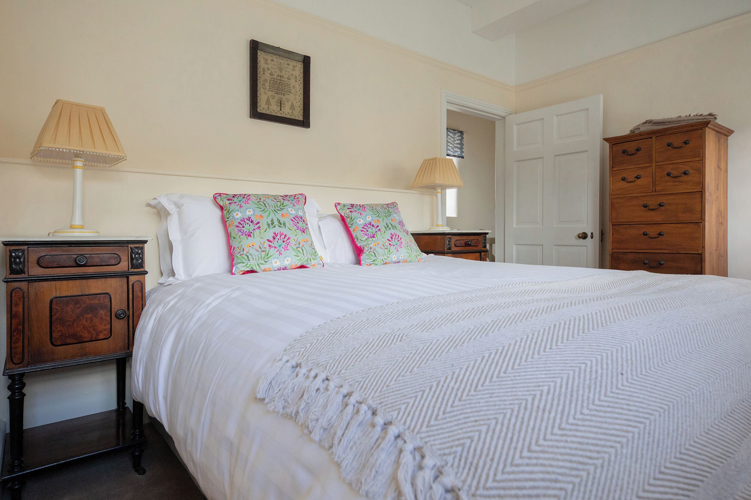 A bedroom with a bed, two bedside tables, a dresser, a door, and a wall-mounted lamp.