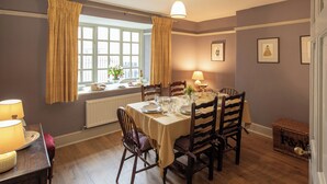 Un comedor con una mesa dispuesta para cuatro, sillas de madera, una ventana con cortinas, una lámpara y cuadros en la pared.