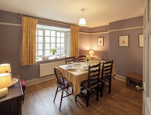 Ein Esszimmer mit Tisch für vier Personen, Holzkstühlen, einem Fenster mit Vorhängen, einer Lampe und gerahmten Bildern an der Wand.