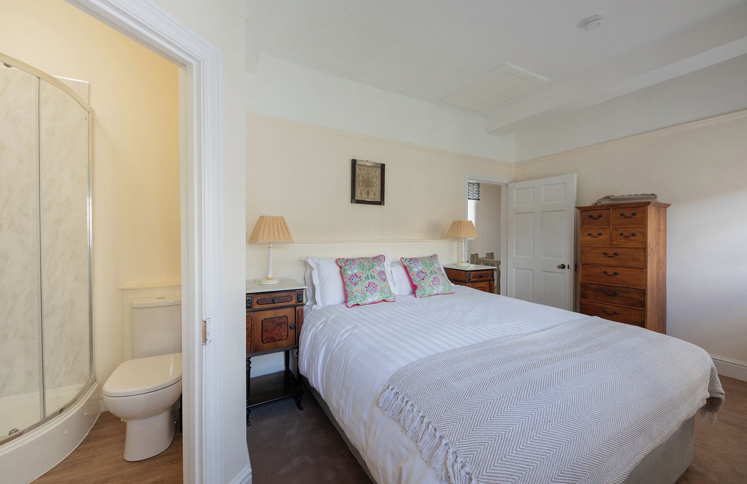 A bedroom with a bed, bedside tables, a wooden dresser, a white door, and a bathroom with a toilet and shower.