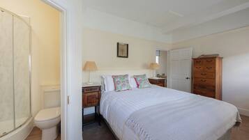 A bedroom with a bed, bedside tables, a wooden dresser, a white door, and a bathroom with a toilet and shower.