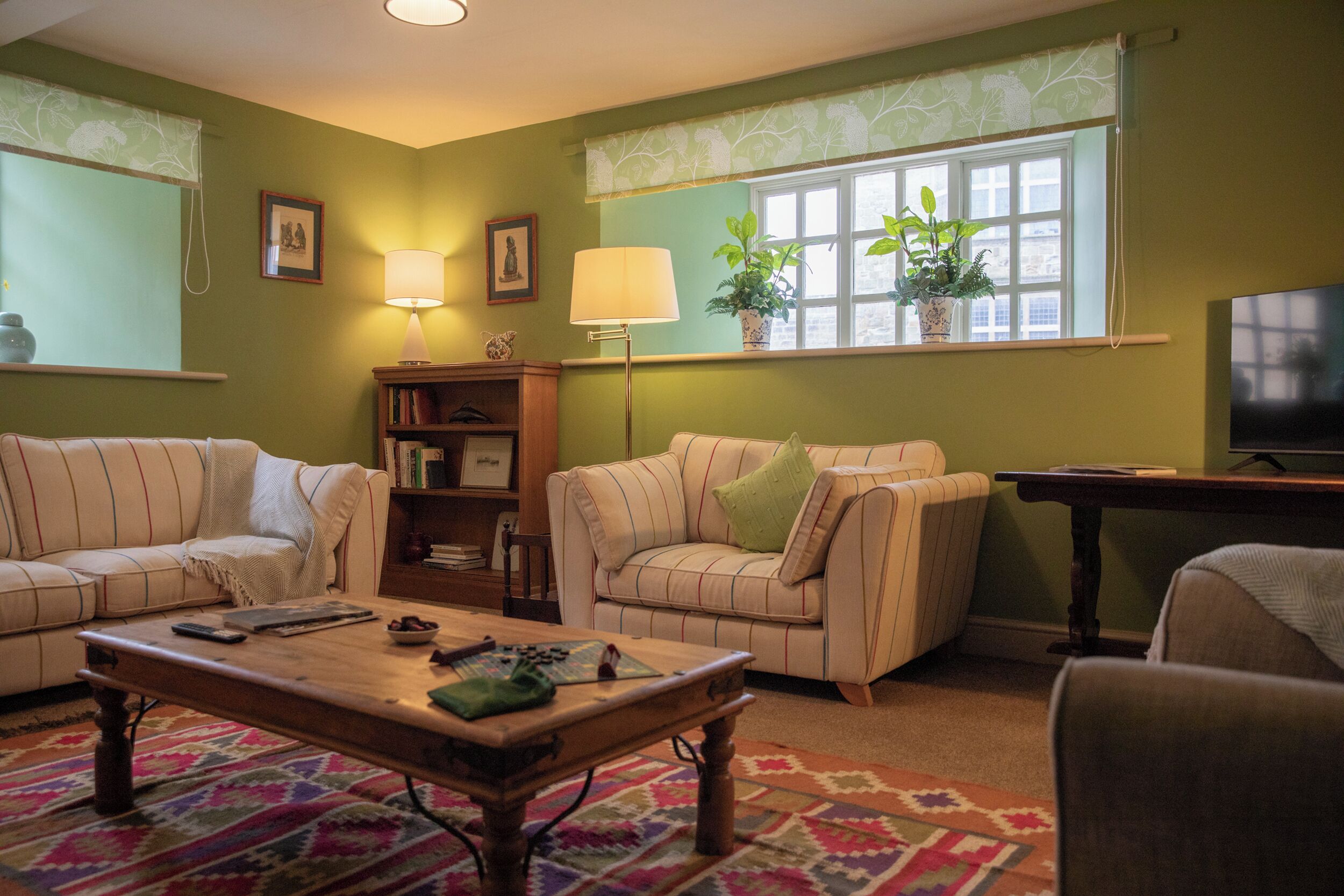 A living room with two sofas, a coffee table, a bookshelf, a TV, and a window with plants.