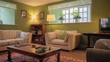 A living room with two sofas, a coffee table, a bookshelf, a TV, and a window with plants.