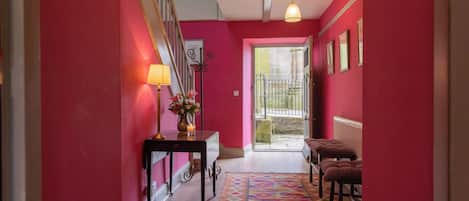 Un pasillo con pared roja, suelo de madera, alfombra estampada, una mesa con lámpara y un banco.