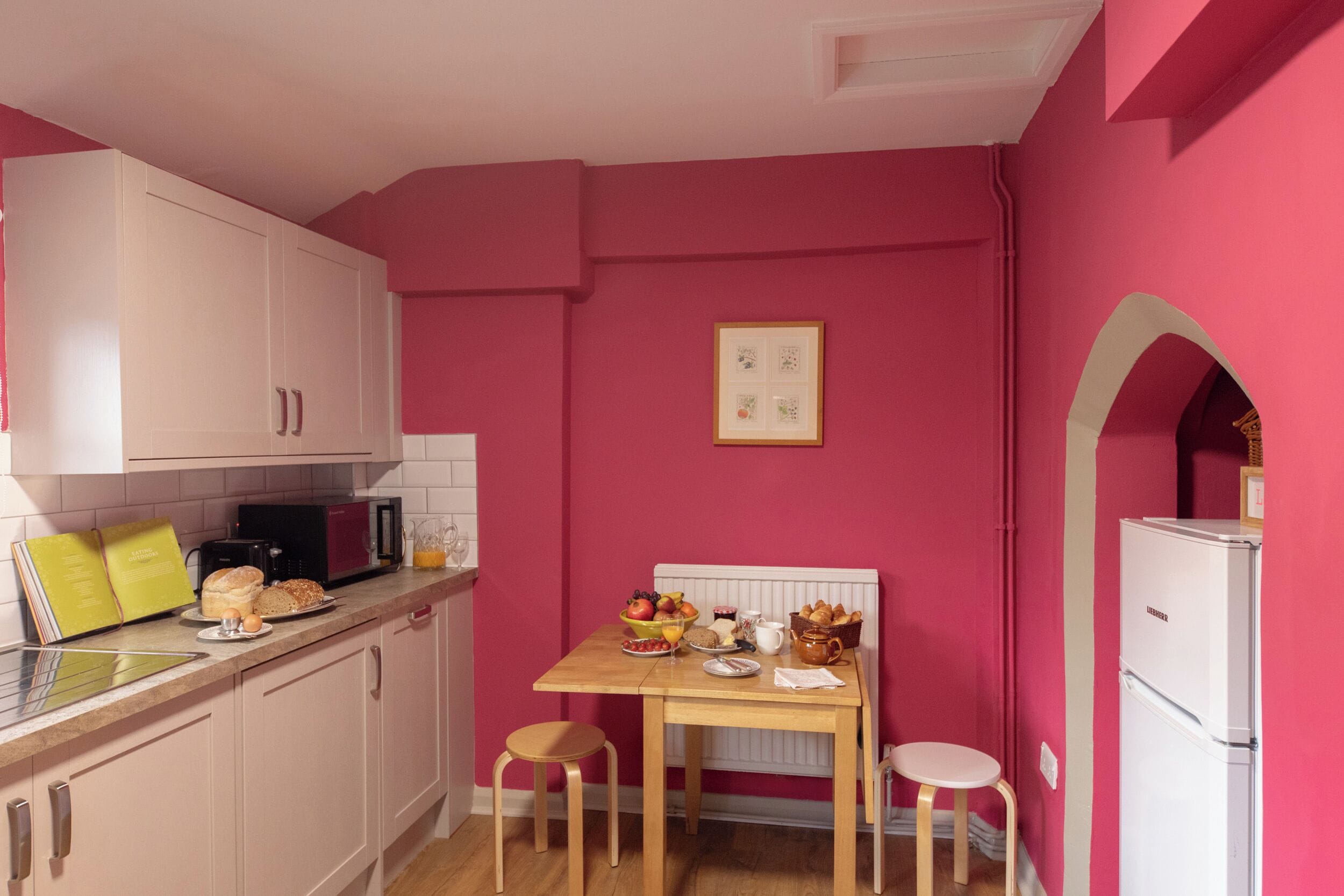 Una cocina con paredes rosadas, gabinetes blancos, una mesa de madera y un refrigerador blanco.