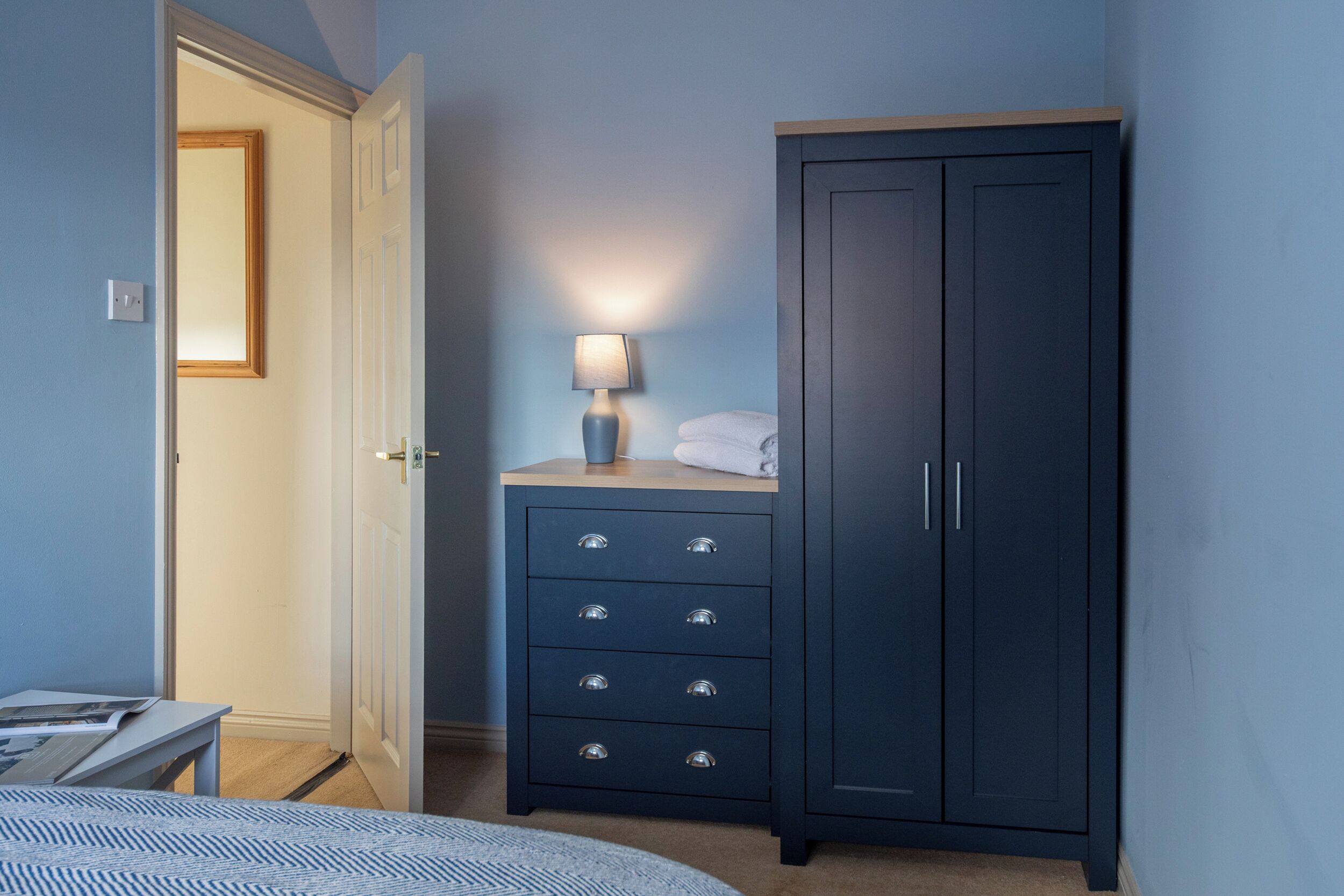Ein Schlafzimmer mit einem Bett, einer Kommode mit Schubladen, einer Lampe und einer Tür zu einem anderen Raum.