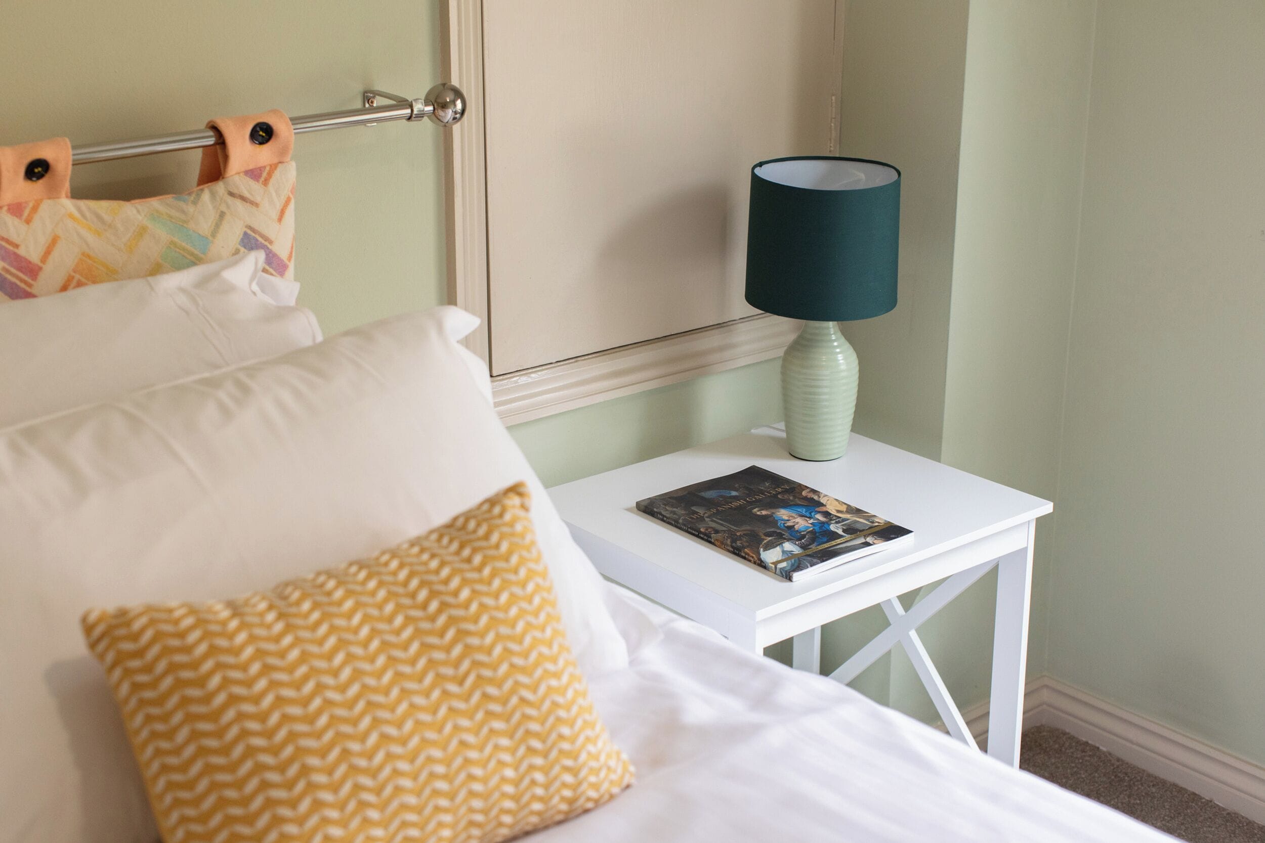 Un letto rifatto con cuscini bianchi e gialli, un comodino bianco con una rivista e una lampada verde.