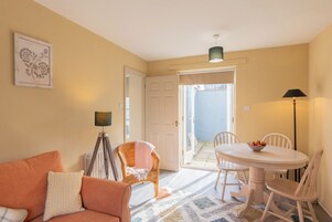 Una habitación con sofá, una mesa de comedor con sillas, una silla de mimbre, una lámpara y una puerta que da a un espacio exterior.