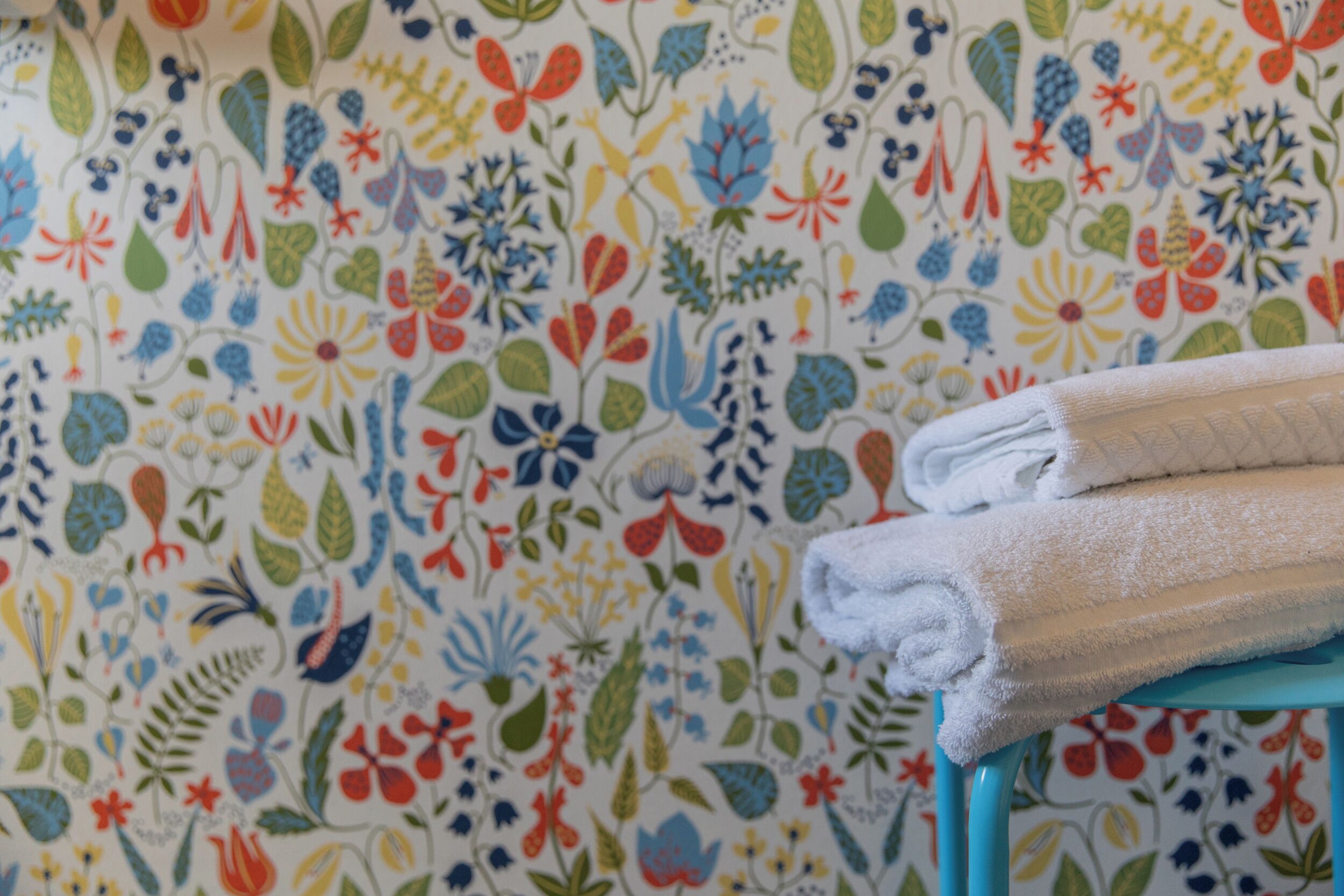 A patterned wallpaper with a floral design, a stack of white towels on a blue chair.