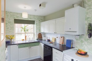 A kitchen with white cabinets, a black dishwasher, a window with a plant, and a fruit bowl on the counter. - Sheriff's Tower Cottage - East (Bishop Auckland)