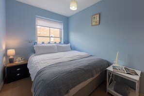 A bedroom with a bed, a nightstand, a bedside table, a window with a view, and a wall-mounted picture. - Sheriff's Tower Cottage - East (Bishop Auckland)