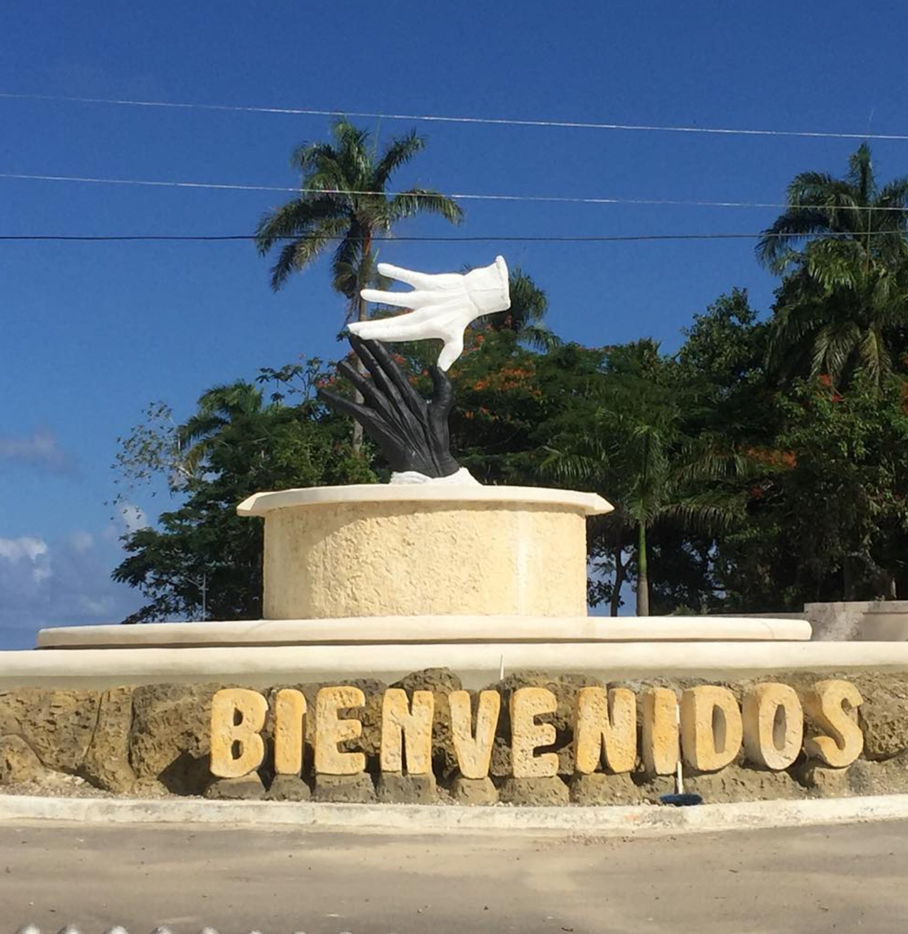 A welcome sign with a hand sculpture and palm trees in the background.