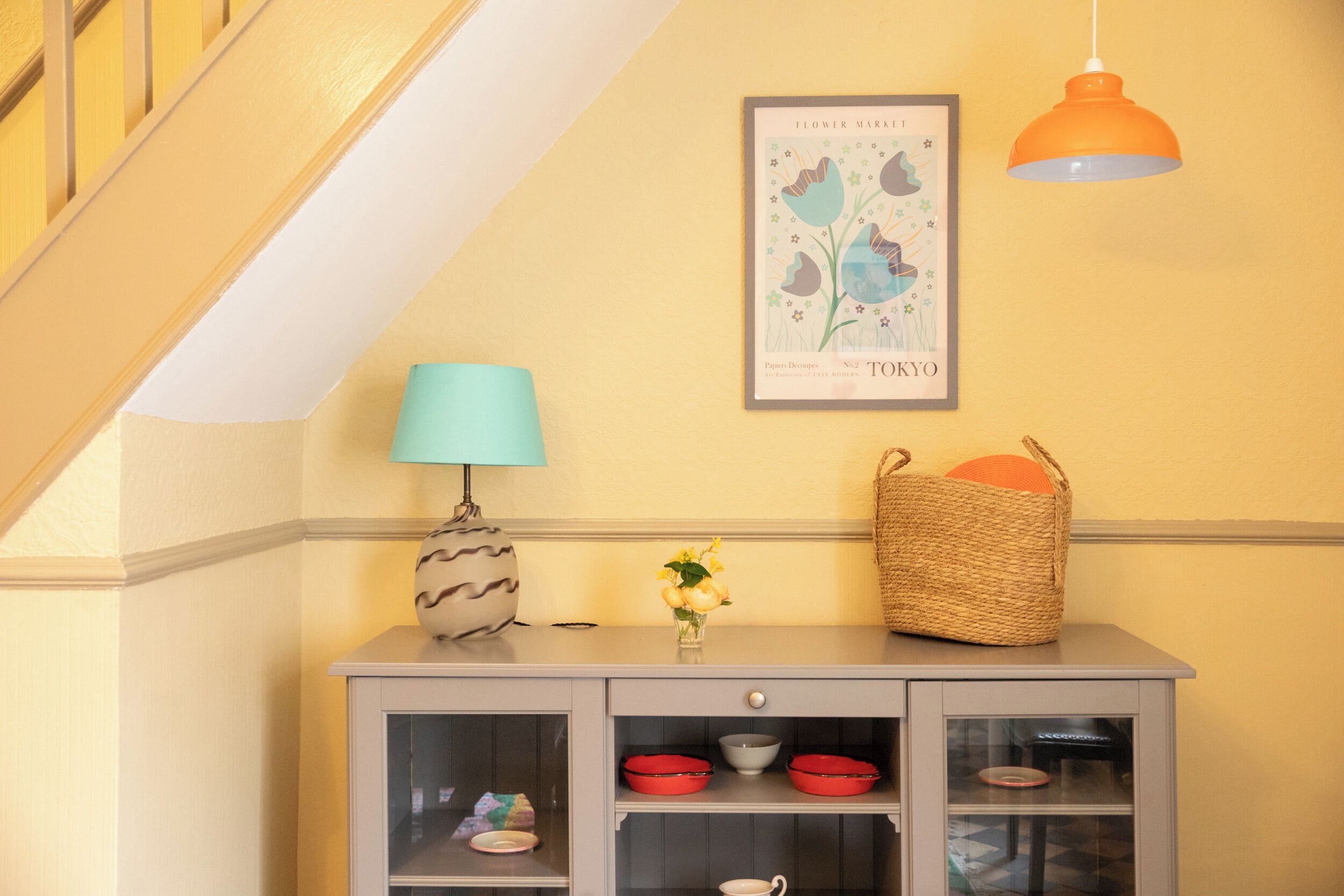 A corner with a grey cabinet, a blue lamp, a vase with yellow flowers, a woven basket, and a framed picture on the wall.