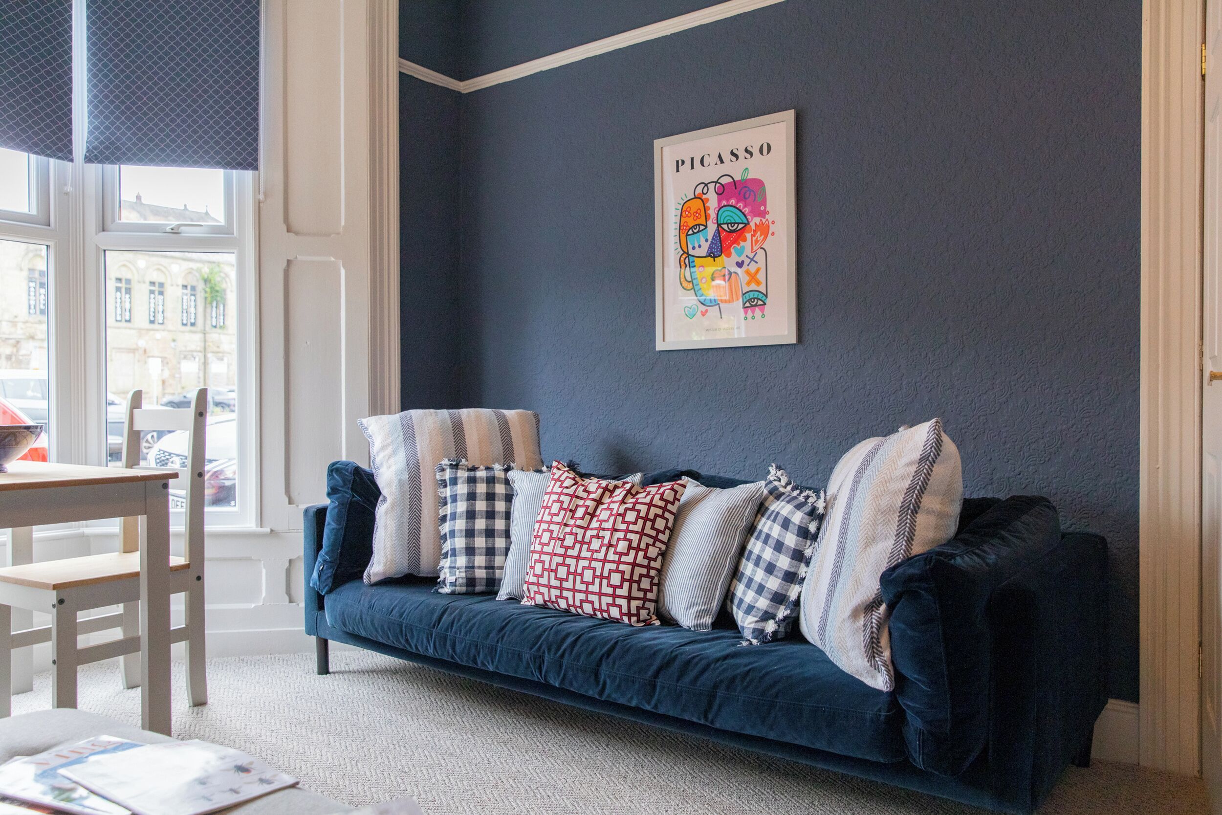 Una habitación con un sofá azul oscuro, cojines estampados, un cuadro enmarcado en la pared y una ventana con vista a la calle.