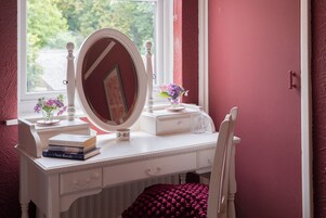 Una tocador blanco con espejo, libros, un florero con flores y una ventana con vistas a la vegetación.