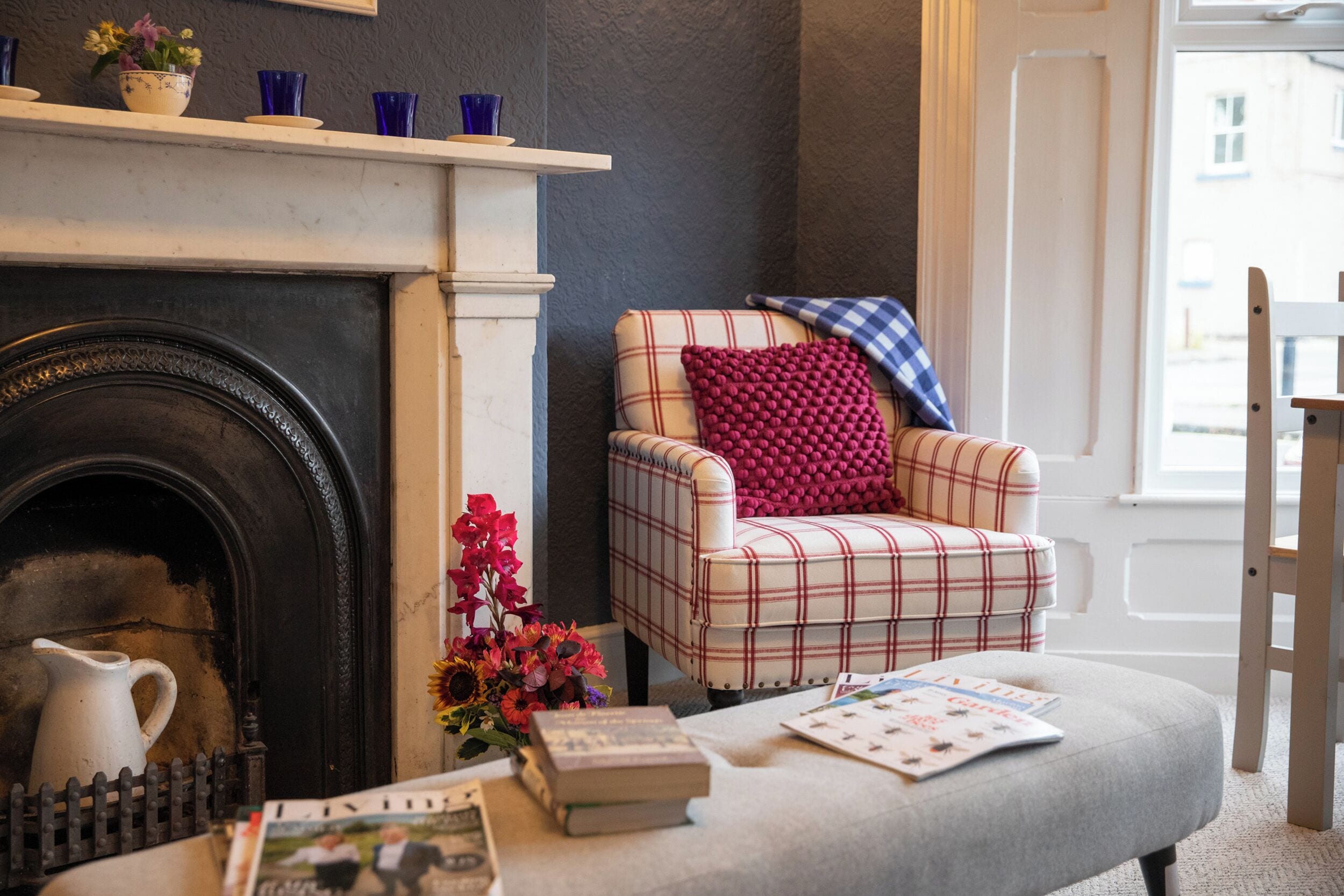 A cozy room with a fireplace, a plaid armchair, a small table with books and a vase of flowers.