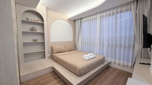 A modern bedroom with a built-in shelf unit, a large bed with a headboard, and a view through sheer curtains. - Relax in Kamala | Rooftop Pool, Gym  | B606 (Tambon Kamala)