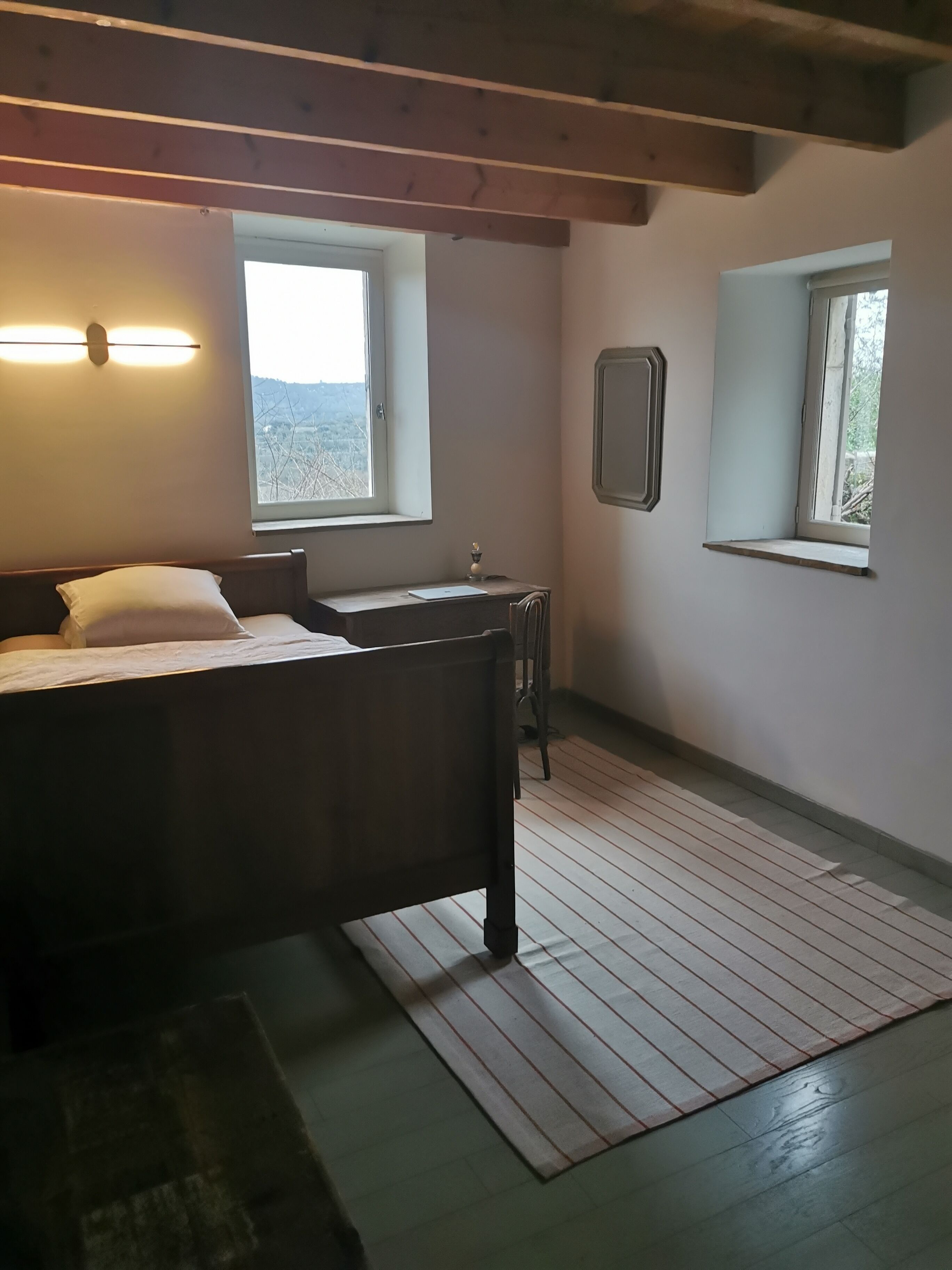 Een slaapkamer met een bed, een bureau, twee ramen en een houten plafond.