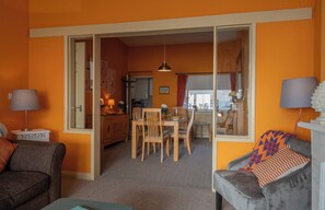 Una sala de estar con sofá, una mesa de comedor con sillas, una cocina al fondo y un ventanal grande.