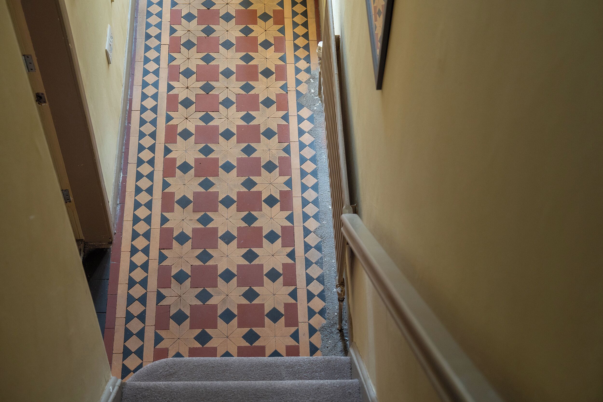 A narrow hallway with patterned floor tiles, a carpeted staircase, and walls with a simple design.