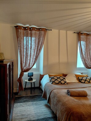Ein Schlafzimmer mit Bett, Vorhängen, einem kleinen Tisch mit Lampe, einem Teppich und einem hölzernen Kleiderschrank.