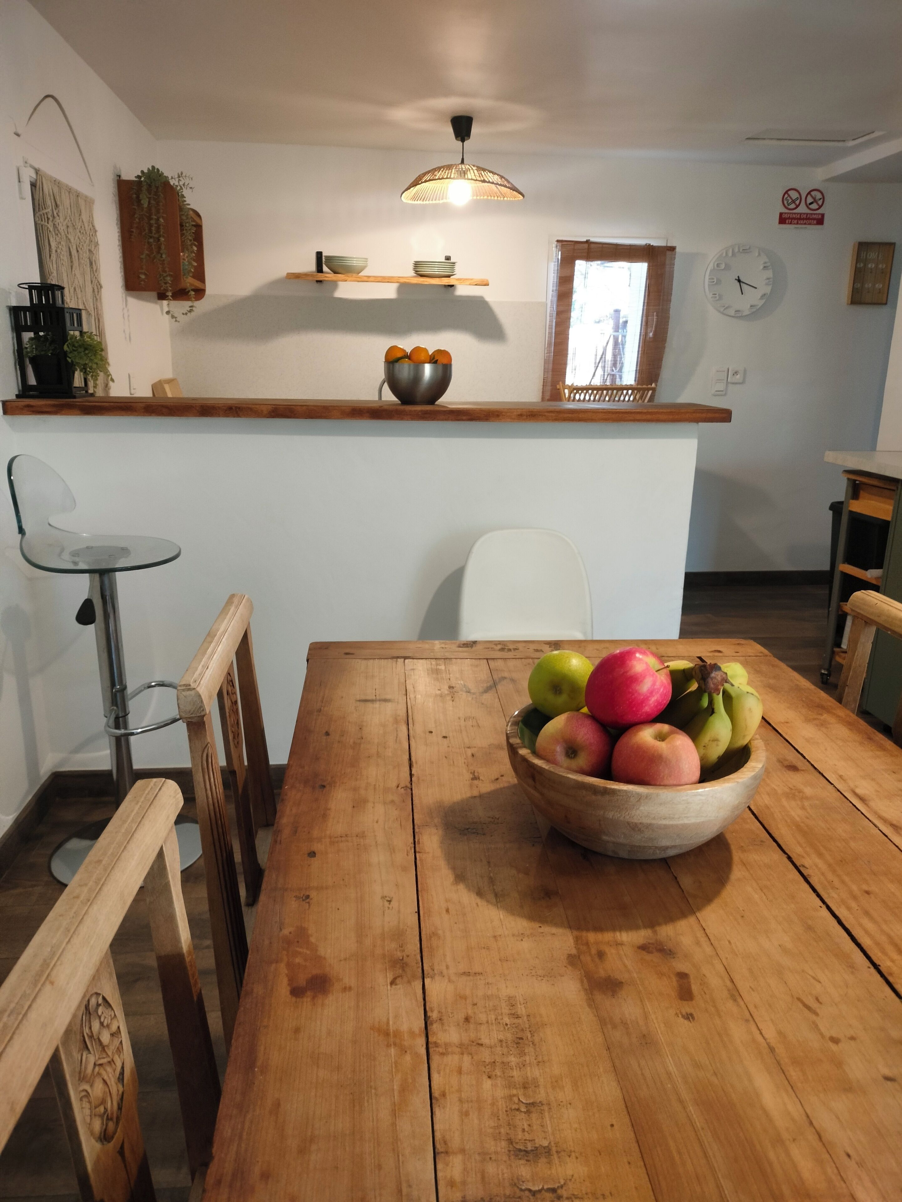 Un comedor con una mesa de madera, sillas, un bol de fruta, una lámpara colgante y una encimera de cocina con estantes.