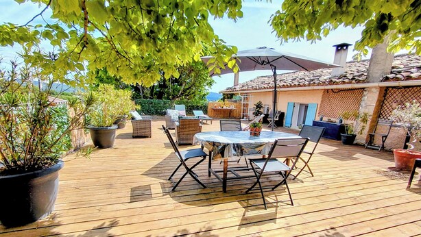 Una terraza de madera con una mesa redonda y sillas, rodeada de plantas en macetas y una pérgola.