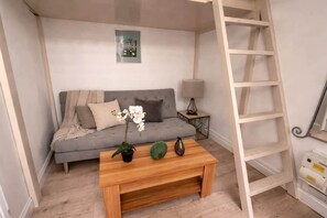 Ein Zimmer mit einem grauen Sofa, einem Holztisch mit Dekoration, einem kleinen Beistelltisch mit Lampe und einer Holzleiter an der Wand.