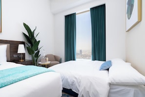 Ein Hotelzimmer mit zwei Betten, einem Nachttisch mit Lampe, einer Zimmerpflanze und Blick auf die Stadt.