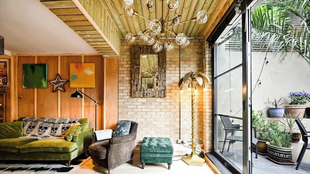 Ein gemütliches Wohnzimmer mit einem grünen Sofa, einem braunen Sessel und einem grünen Hocker. An der Wand befindet sich ein großer Spiegel, eine Chandelieber und vor dem Fenster stehen Blumentöpfe.